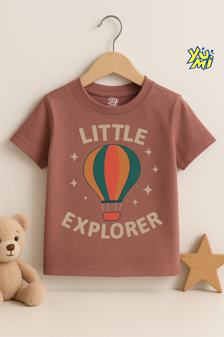 Children's t-shirt with 'Little Explorer' design on a hanger, surrounded by a teddy bear and star decorations.